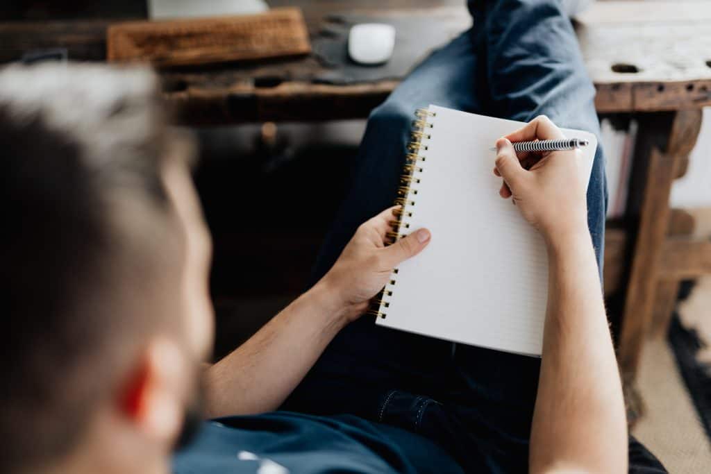 A man dressed in casual blue jeans making notes in a spiral bound notebook. He is using a biro decorated with white rings down its length and has his feet up on a workbench.