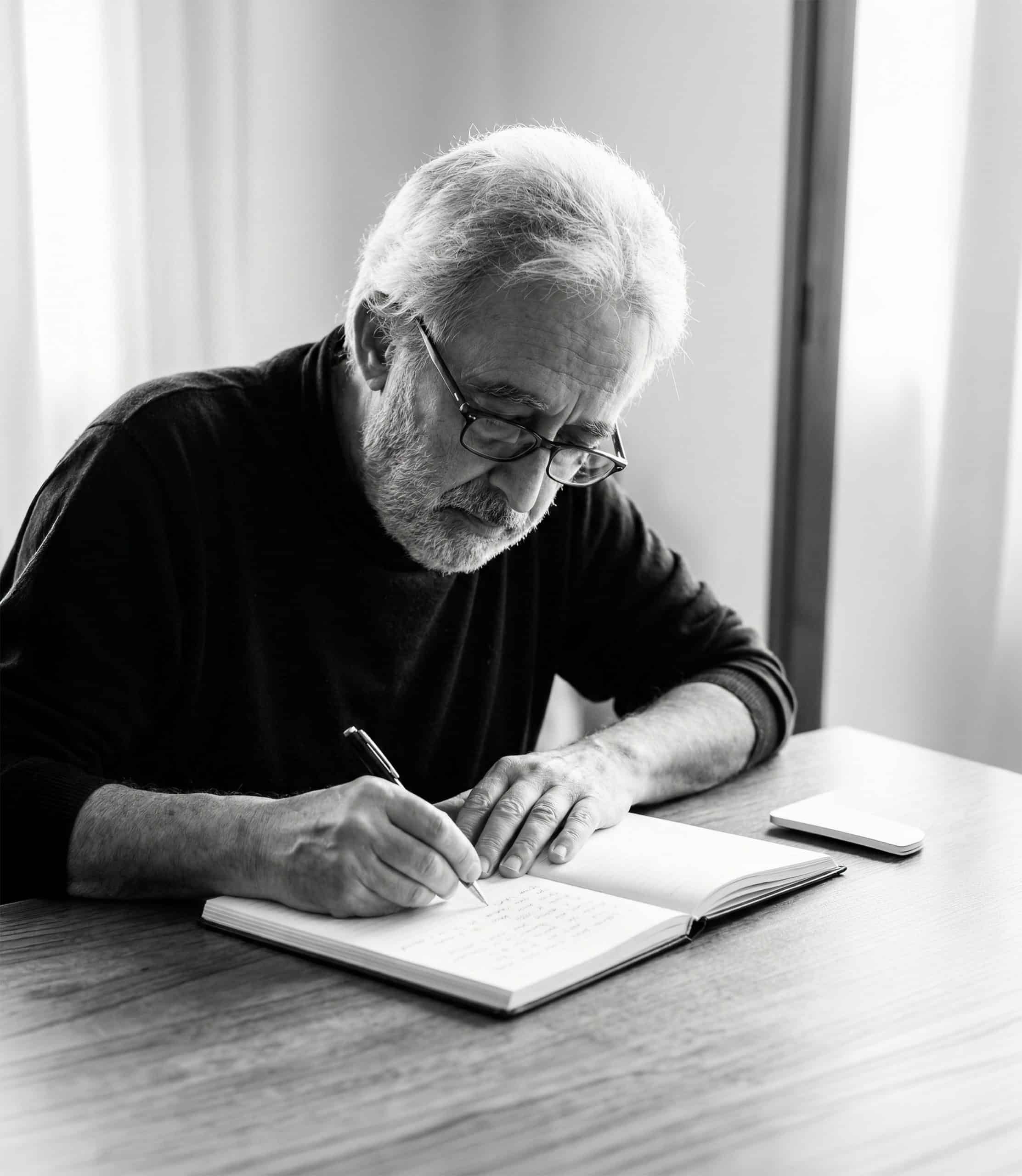 A white haired but fit looking older man who has his jumper sleeves rolled up while writing with a fountain pen in a book with empty pages.